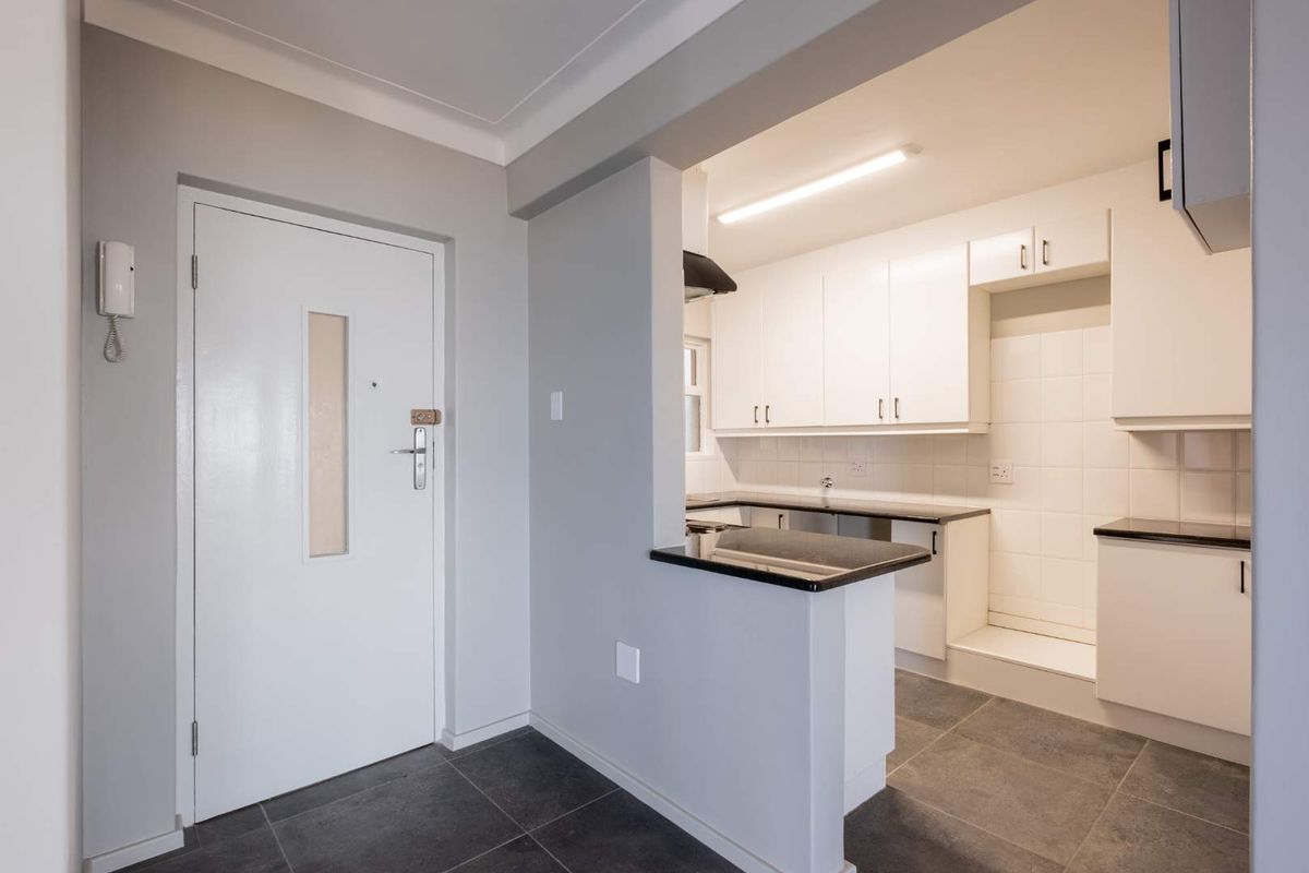 A clean, modern kitchen featuring white cabinetry, dark countertops, tiled backsplash, and grey floor tiles, with an adjacent entrance door and intercom system.