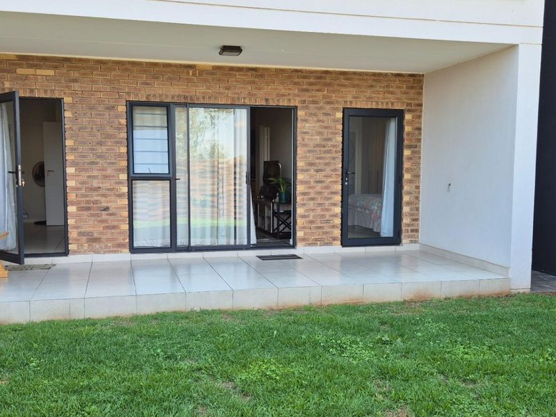 A covered patio area featuring tiled flooring, exposed brickwork, and multiple glass access points leading into the apartment, overlooking a private garden space.