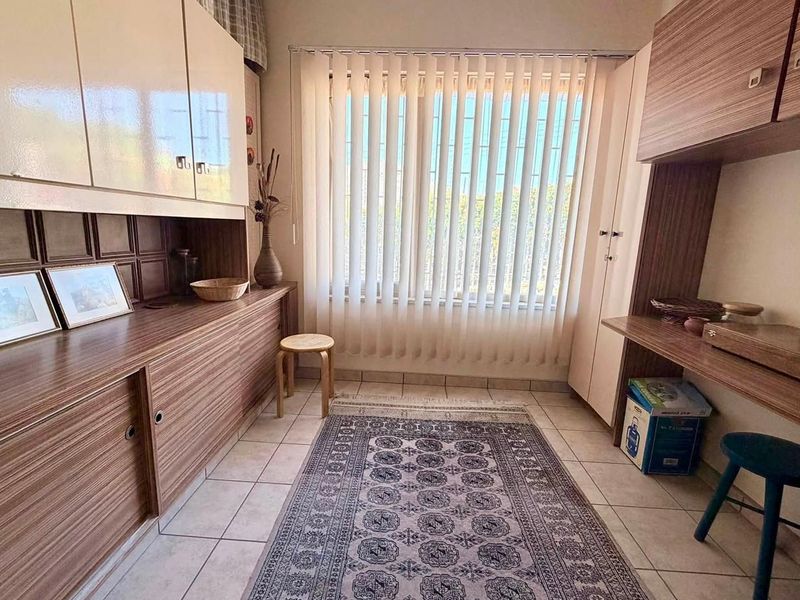A functional home office or study area featuring extensive built-in cabinetry, tiled flooring, and large windows with vertical blinds.