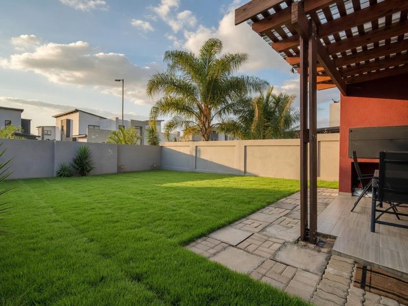 A well-maintained rear garden featuring a paved patio area with a wooden pergola and a built-in braai, enclosed by a solid boundary wall.