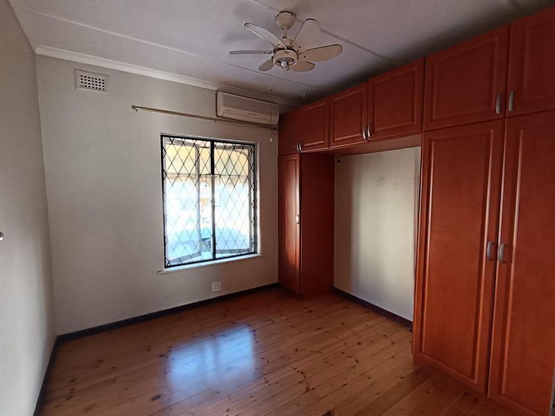 A spacious bedroom featuring polished timber flooring, built-in wooden cabinetry, a ceiling fan, and an air conditioning unit.