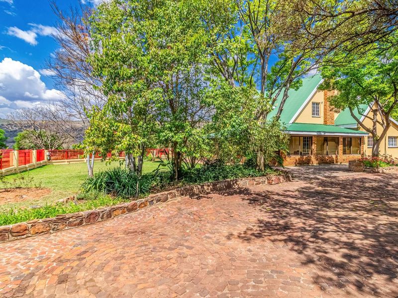 A charming farm-style residence featuring a cobblestone driveway, established trees, and a covered veranda, set within a well-maintained garden with scenic landscape views.