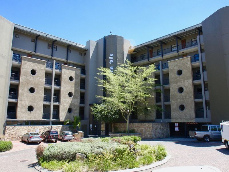 Front elevation of The Falls apartment complex featuring modern architectural design with circular windows, stone cladding, and a paved driveway entrance.