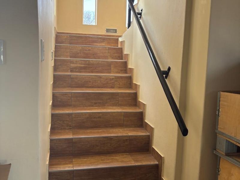 A bright staircase featuring wood-look tiled steps and a modern black metal handrail leading to an upper level.