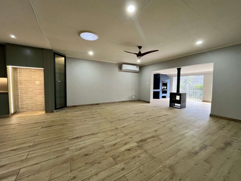 Spacious open-plan living area featuring wood-look tile flooring, modern recessed lighting, a built-in fireplace, and an integrated braai area leading to the patio.