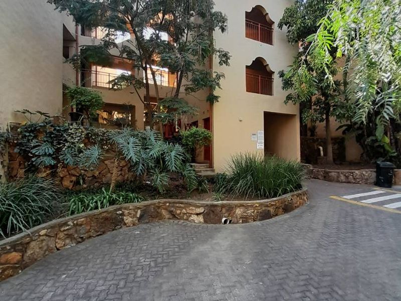 Exterior view of an apartment building featuring a paved driveway, stone retaining walls, and established garden landscaping with balconies.