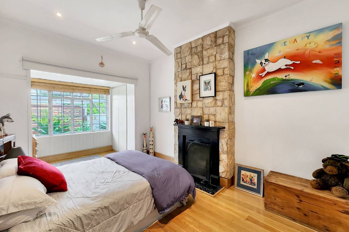 A bright bedroom featuring hardwood flooring, a stone-clad fireplace, ceiling fan, and built-in storage with large windows overlooking the garden.