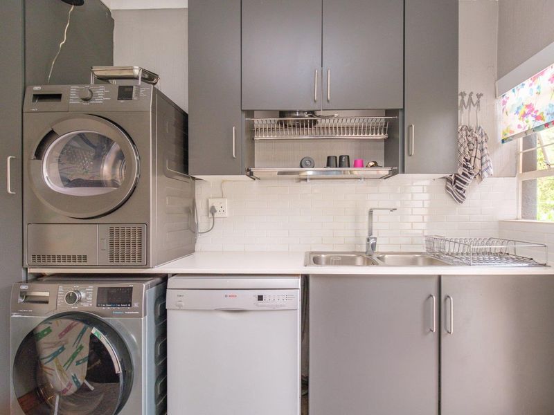 A functional laundry area featuring modern grey cabinetry, a stainless steel sink, subway tile backsplash, and space for stacked laundry appliances and a dishwasher.