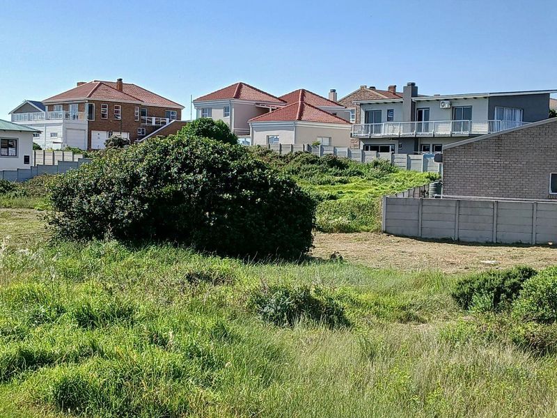 A view of an undeveloped residential plot featuring overgrown grass and a precast concrete boundary wall, surrounded by established neighboring homes.