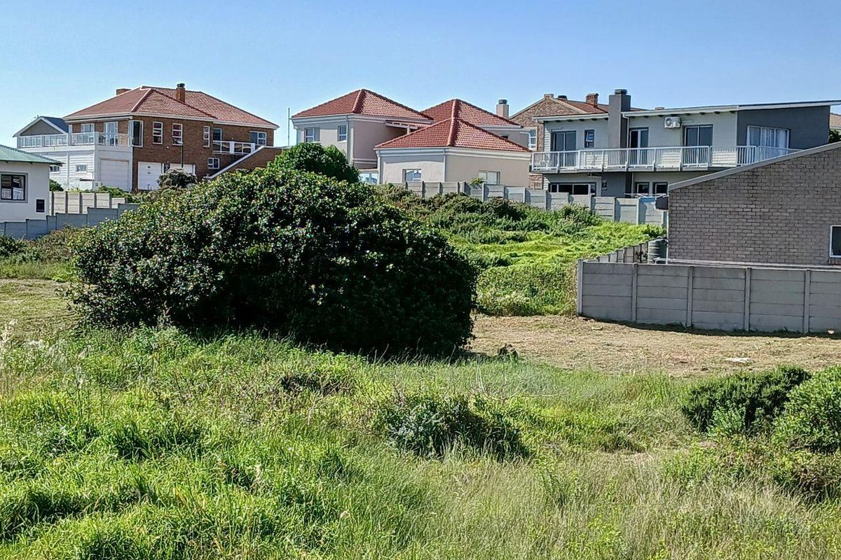 A view of an undeveloped residential plot featuring overgrown grass and a precast concrete boundary wall, surrounded by established neighboring homes.