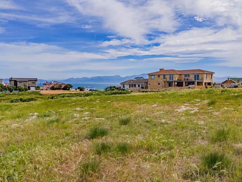 A wide-angle view of a vacant residential plot featuring natural coastal vegetation with surrounding neighborhood homes and distant mountain scenery.