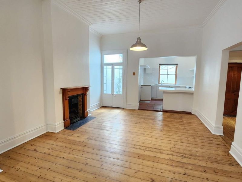 An open-plan living area featuring original timber flooring, a classic fireplace with a timber mantel, and a connected kitchen space with a breakfast bar.