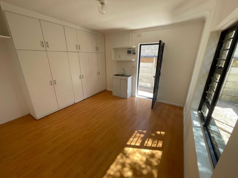 A bright, empty bedroom featuring extensive built-in cupboards, laminate flooring, and a small kitchenette sink area with direct access to an outdoor space.