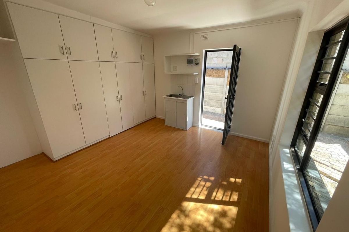 A bright, empty bedroom featuring extensive built-in cupboards, laminate flooring, and a small kitchenette sink area with direct access to an outdoor space.