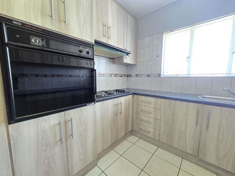 A functional kitchen featuring light wood-look cabinetry, dark laminate countertops, a built-in oven, hob, and tiled flooring with ample natural light.