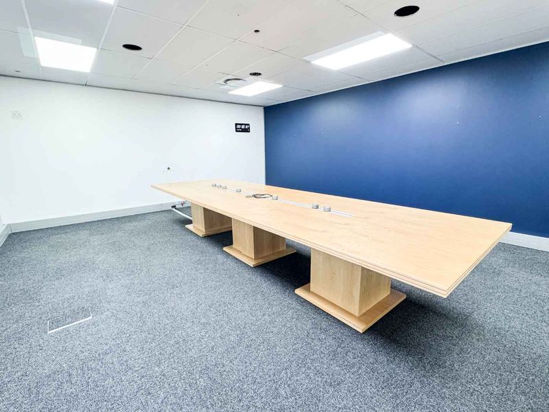 A professional office boardroom featuring a large built-in table, carpeted flooring, and a modern suspended ceiling with recessed lighting.