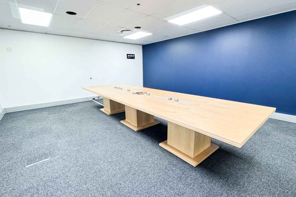 A professional office boardroom featuring a large built-in table, carpeted flooring, and a modern suspended ceiling with recessed lighting.