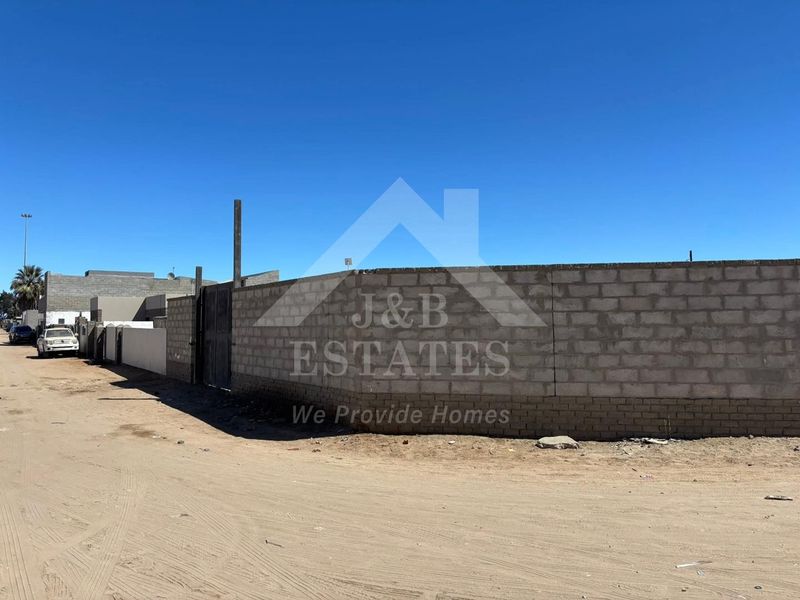 A view of the vacant residential plot featuring a perimeter block wall and gate, situated along an unpaved street.