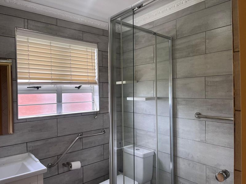 A modern bathroom featuring grey ceramic wall tiles, a glass shower enclosure, and installed safety grab rails for accessibility.