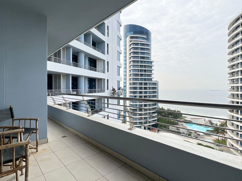 A spacious covered balcony featuring tiled flooring and a stainless steel railing, offering expansive views of the surrounding high-rise buildings and coastal landscape.