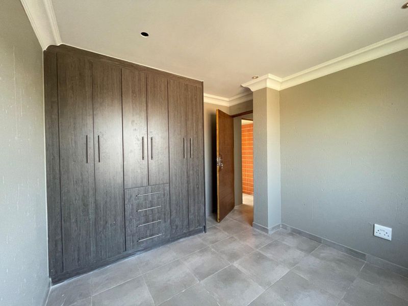 A clean, empty bedroom featuring modern dark wood built-in cupboards, neutral grey wall paint, and tiled flooring with decorative ceiling cornicing.