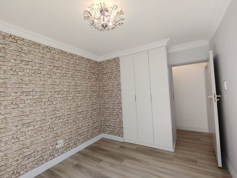 A bright, empty bedroom featuring modern laminate flooring, built-in cupboards, and a decorative ceiling light fixture with a feature brick-effect wall.