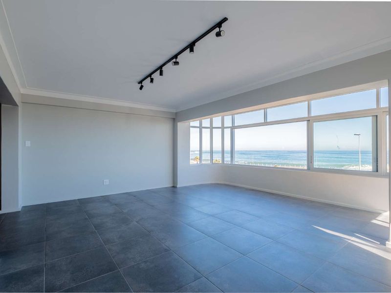 A bright, spacious, and empty living area featuring modern dark tiled floors, track lighting, and expansive windows offering panoramic ocean views.