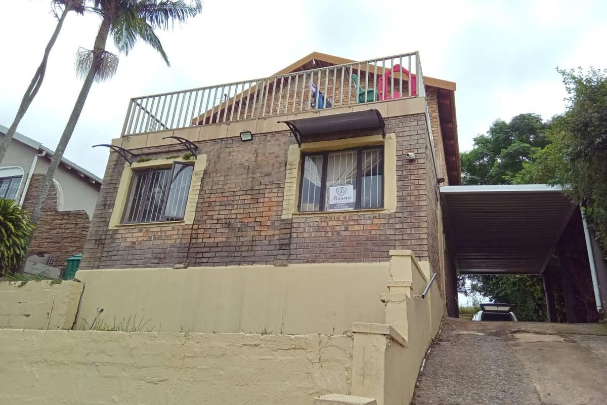 Front elevation of a brick house featuring a balcony, carport, and concrete driveway, showing signs of wear on the exterior finishes and retaining wall.