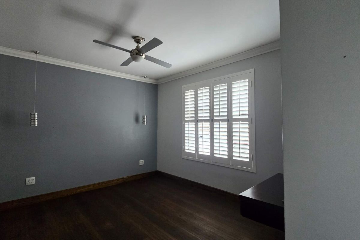 A spacious, empty bedroom featuring dark hardwood flooring, crown molding, a ceiling fan, and white plantation shutters.