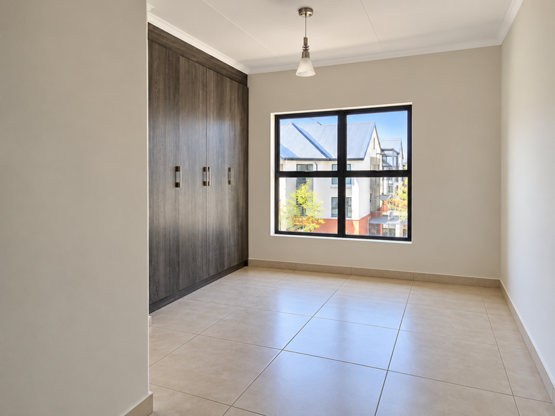 A bright, empty bedroom featuring neutral tiled flooring, modern built-in cupboards, and a large window providing natural light.