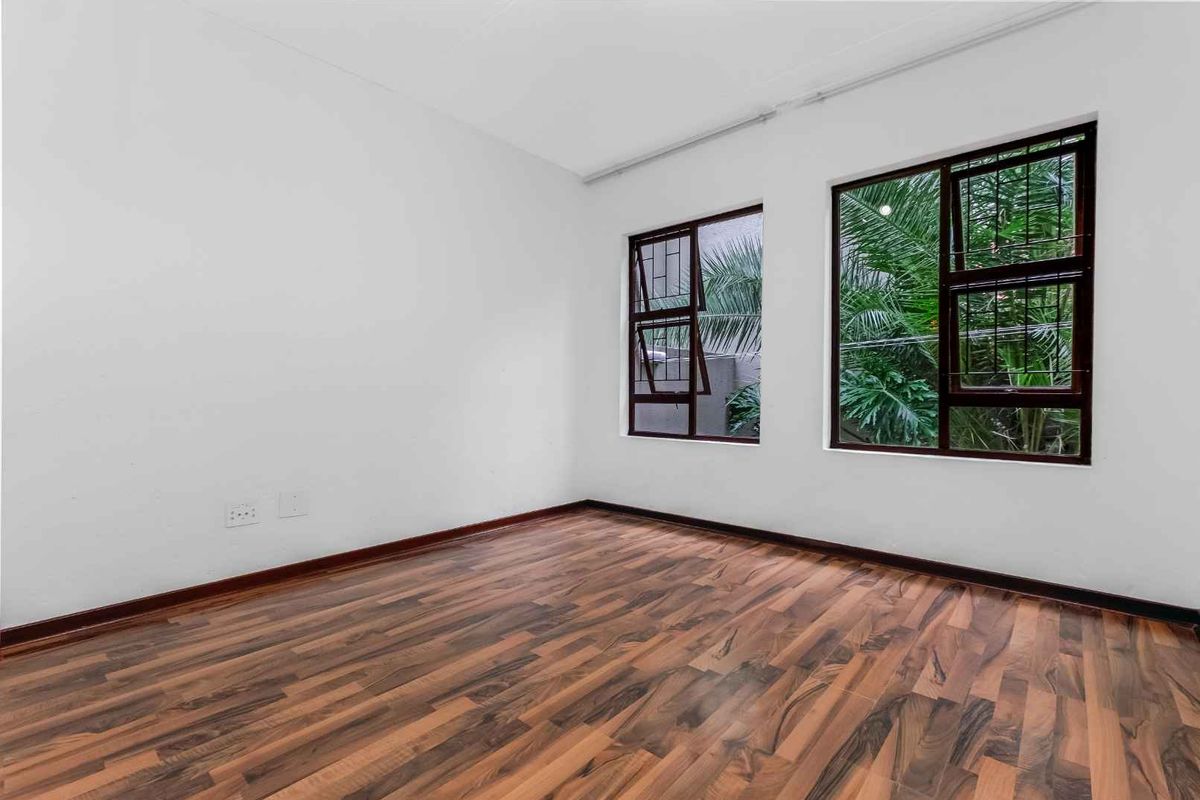 A bright, empty bedroom featuring dark wood-look laminate flooring and large windows with dark frames overlooking greenery.