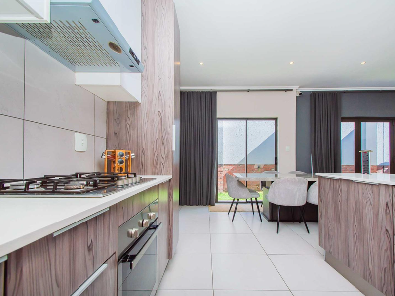 Modern open-plan kitchen featuring wood-grain cabinetry, stone countertops, integrated gas hob, and oven, flowing seamlessly into a dining area with access to the patio.