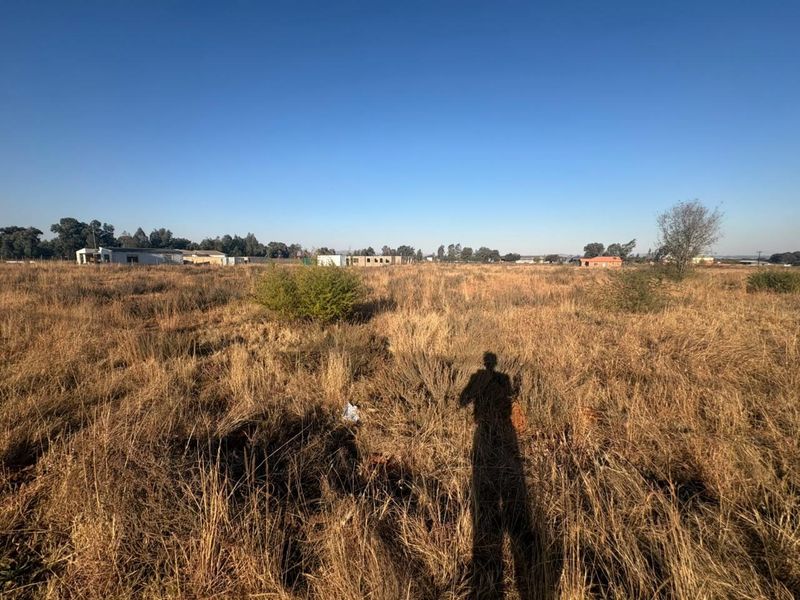 A wide-angle view of a vacant residential plot featuring dry grass and surrounding neighborhood development in the distance.