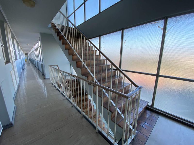 A bright, open apartment building hallway featuring a central staircase with metal balustrades and large windows providing natural light.