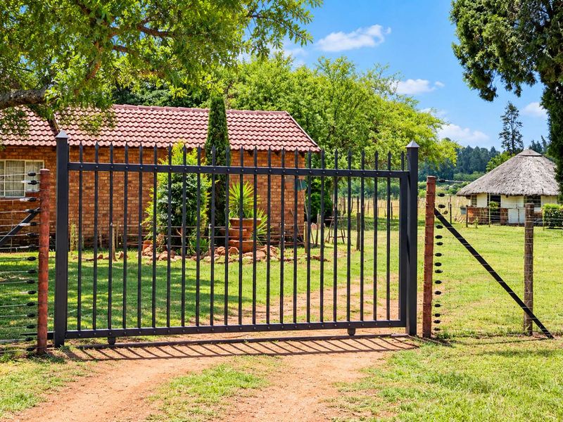 A secure entrance featuring a metal gate with electric fencing, leading to a property with brick and thatched roof buildings on a small holding.