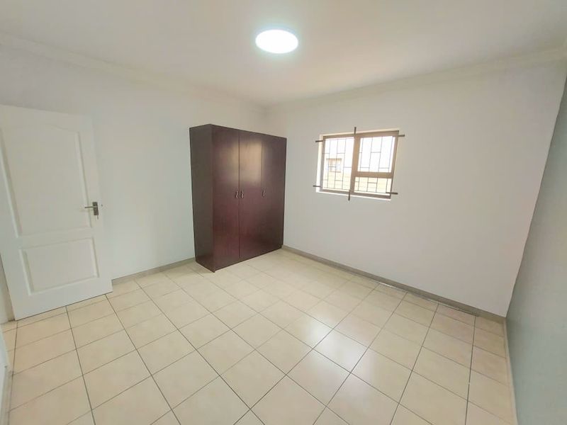 A clean, empty bedroom featuring neutral tiled flooring, a built-in wardrobe, and a window fitted with security bars.