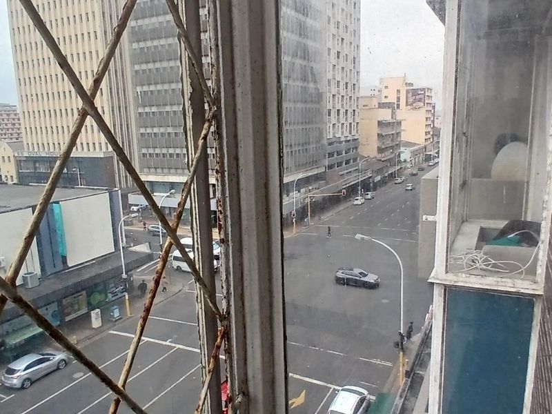 An elevated urban view from an apartment window overlooking a city street intersection, featuring visible exterior brickwork and weathered security bars.