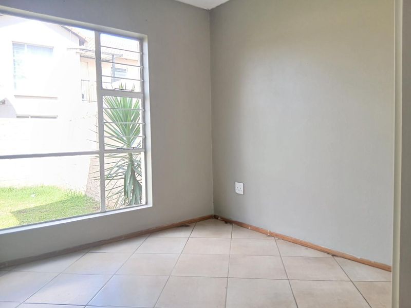 A bright, empty bedroom featuring neutral wall paint, tiled flooring, and a large window with security bars overlooking the garden area.