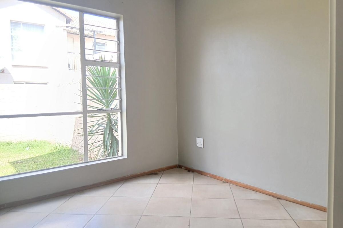 A bright, empty bedroom featuring neutral wall paint, tiled flooring, and a large window with security bars overlooking the garden area.