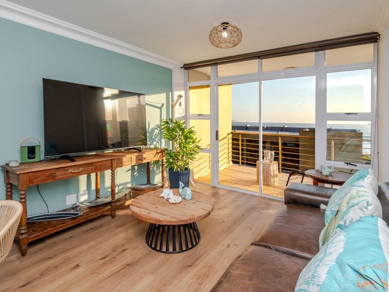 A bright living room featuring laminate flooring, decorative ceiling cornice, and large sliding glass doors that open onto a balcony with ocean views.