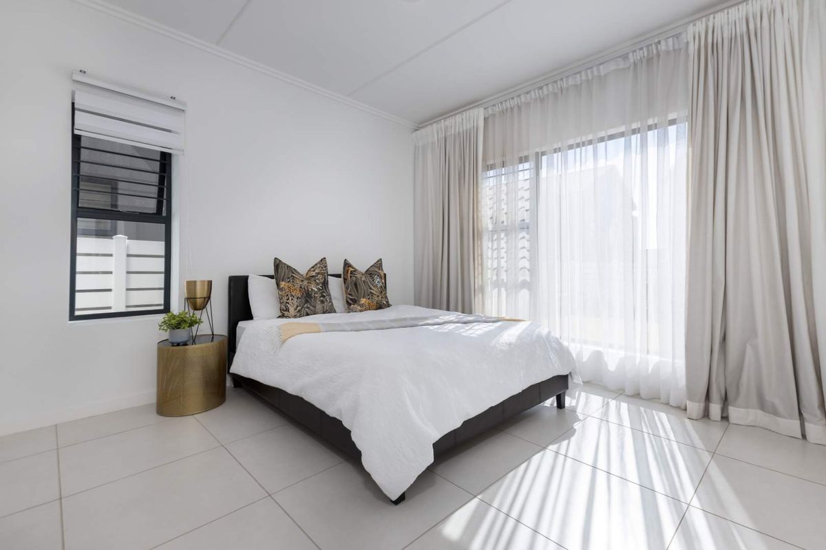 A bright and modern bedroom featuring large sliding glass doors with floor-to-ceiling curtains, tiled flooring, and a window with adjustable louvers.