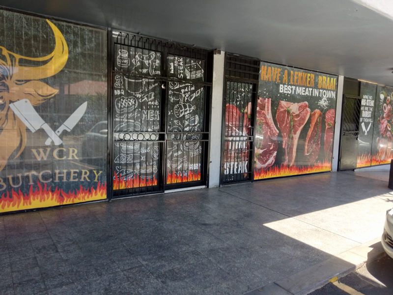 Front facade of a retail butchery featuring large glass display windows, security gates, and a covered tiled walkway entrance.