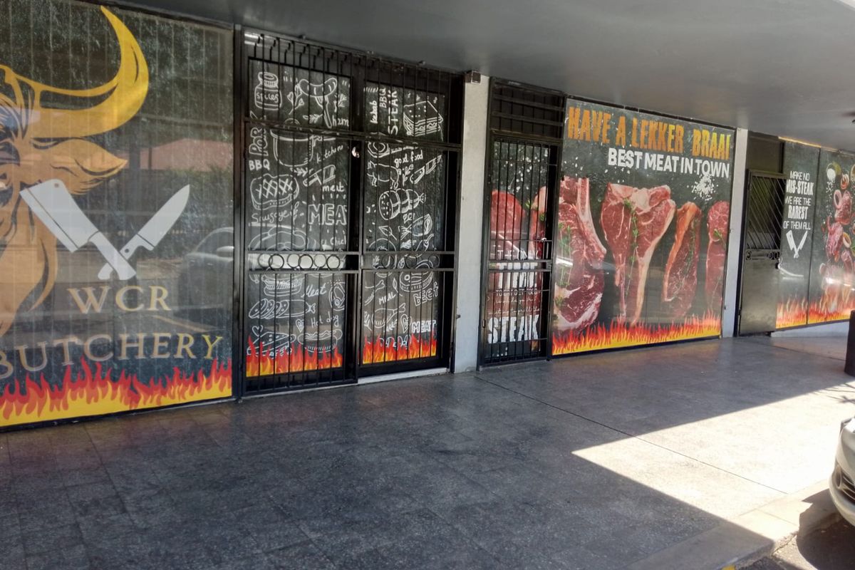 Front facade of a retail butchery featuring large glass display windows, security gates, and a covered tiled walkway entrance.