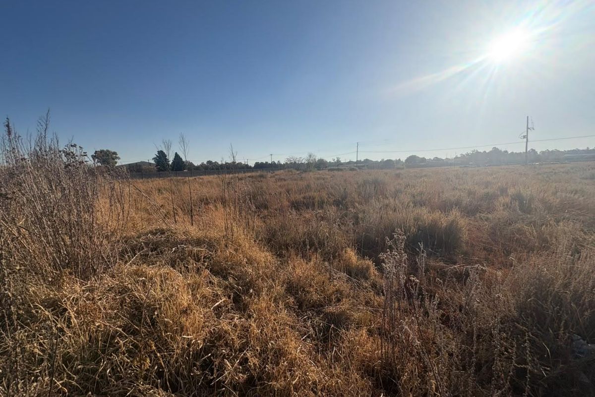 A wide-angle view of a vacant residential plot featuring dry grass and perimeter fencing under a clear blue sky.