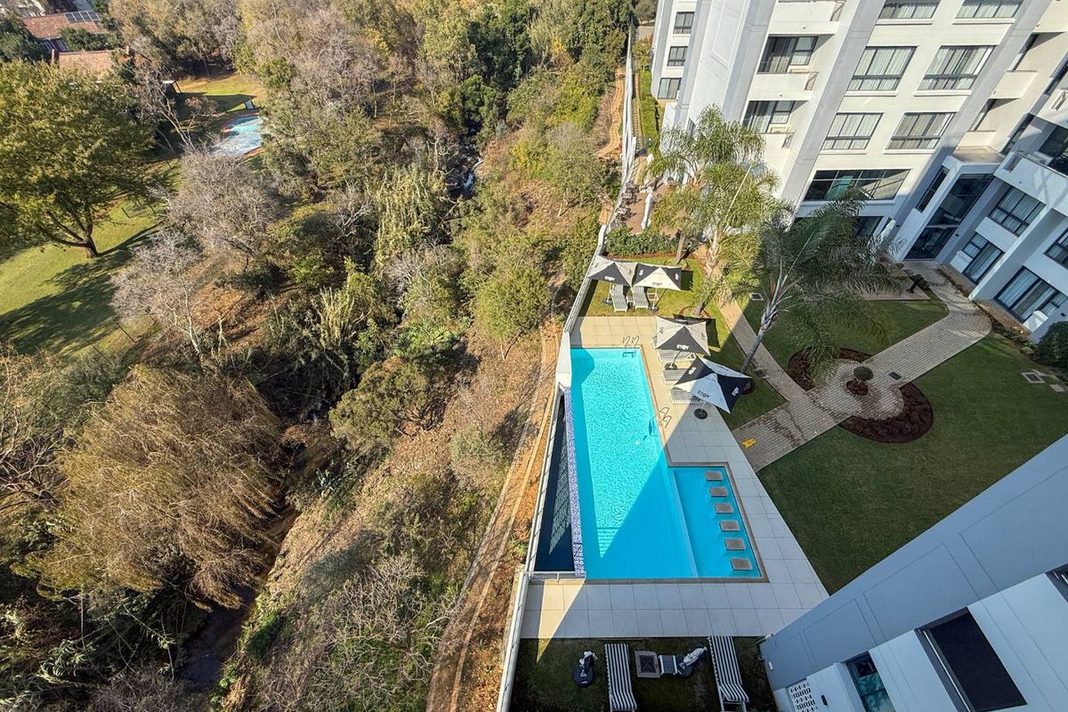 Aerial view of the apartment complex featuring a well-maintained swimming pool, paved sun deck, and landscaped garden areas adjacent to a natural wooded ravine.