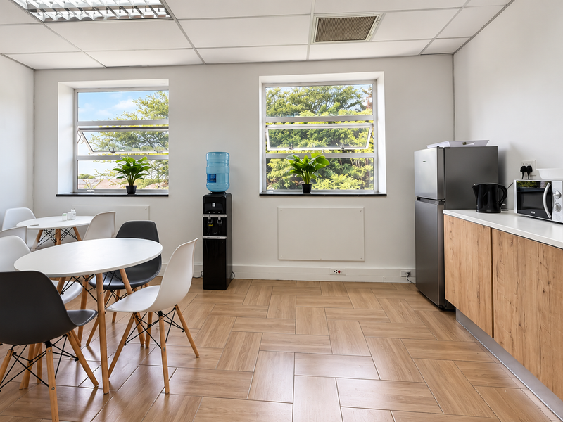 A bright office kitchenette featuring wood-look tile flooring, modern cabinetry, and ample natural light from dual windows.