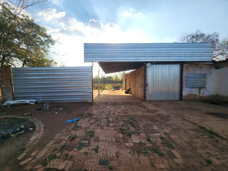 Rear exterior view of a mixed-use property featuring corrugated metal fencing, a covered structure with a metal roof, and brick paved ground area.