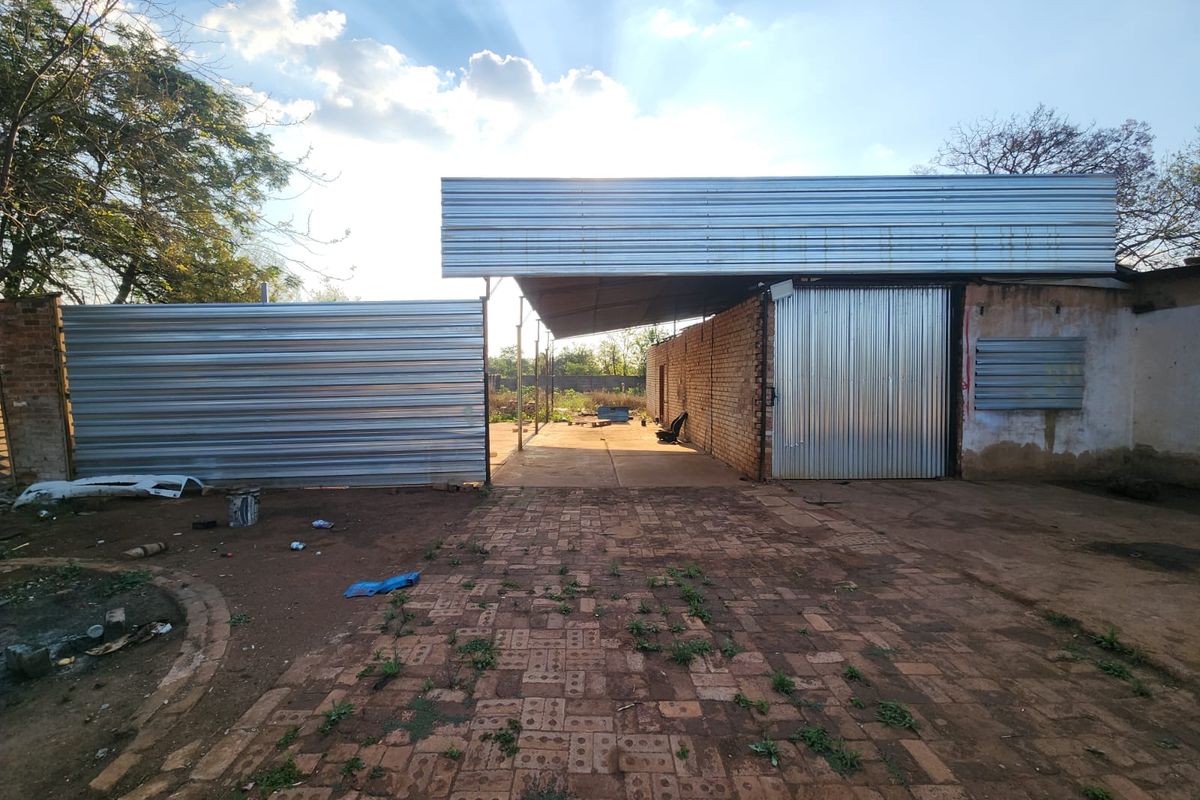 Rear exterior view of a mixed-use property featuring corrugated metal fencing, a covered structure with a metal roof, and brick paved ground area.