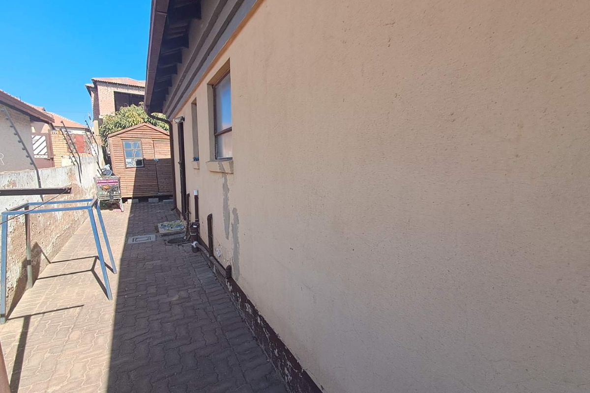 Side exterior view of the house featuring paved walkway, exterior wall with visible wear, and a small wooden storage shed in the rear.