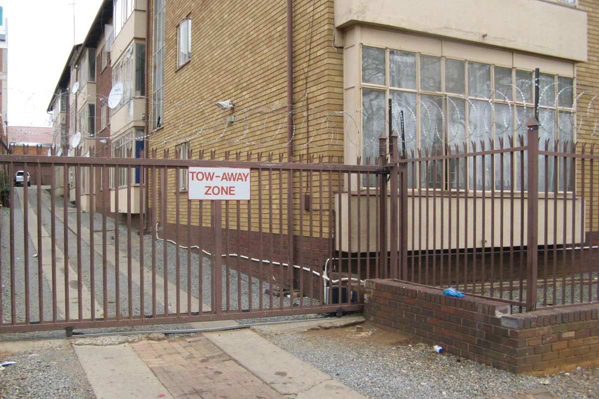 Exterior view of an apartment building featuring a brick facade, metal security gate, and perimeter security fencing with razor wire.